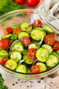 a glass bowl filled with cucumbers and tomatoes on top of a table next to broccoli