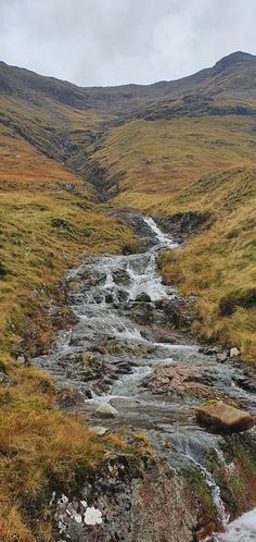 a small stream running through a lush green hillside covered in grass and rocks with mountains in the background