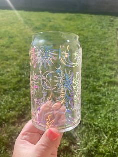 a hand holding a glass with flowers painted on it in front of some green grass