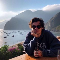 a man sitting at a table with a beer in his hand and boats in the water behind him