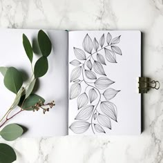 an open notebook on top of a table next to a leafy plant and a pen