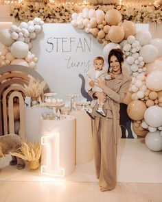 a woman holding a baby standing in front of a backdrop with balloons and streamers