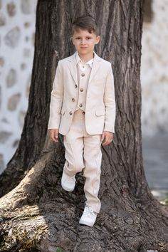 a young boy in a suit standing next to a tree with his hands in his pockets