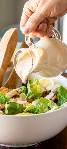 a person pouring dressing into a salad in a white bowl on a wooden table next to bread