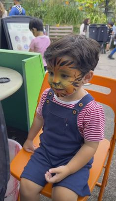 a young boy with face paint sitting on an orange chair in front of a table