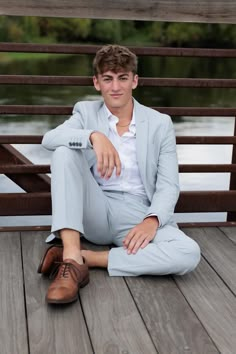 a young man in a suit and shoes sitting on a wooden deck next to a body of water