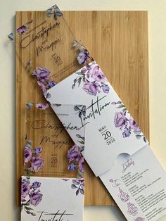 the wedding stationery is laid out on a cutting board with purple flowers and greenery