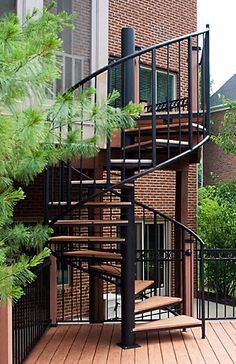 a wooden deck with a spiral staircase next to a brick building and green trees in the foreground