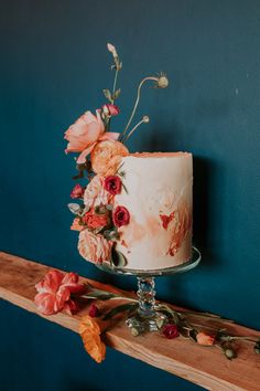 a white cake with pink flowers on top sitting on a wooden shelf next to a blue wall