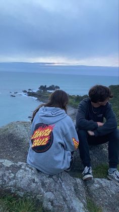 two people sitting on top of a rock next to the ocean and looking at the water