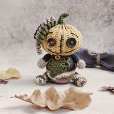 a small crocheted doll sitting on top of a leaf covered floor next to leaves