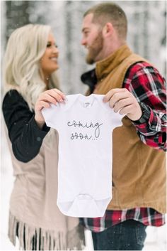 a man and woman holding up a baby's t - shirt in the snow