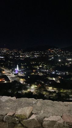 a person sitting on top of a rock wall looking at the city lights in the distance