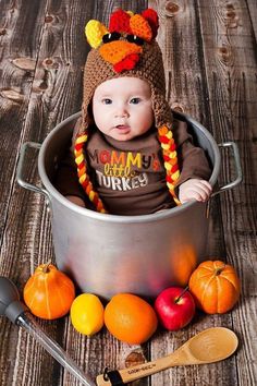 a baby wearing a turkey hat sitting in a pot with apples and oranges next to it