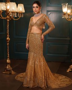 Metallic gold fishcut lehenga with all-over sequin, crystal, and cutdana embroidery with a flowing trail at the back. Paired with a delicate full sleeves blouse with hanging tassel detailing and embroidered dupatta in net.DELIVERY TIMEPlease allow 8-12 weeks for your outfit to arrive.FABRIC DETAILSNetProfessional cleaning only. Glamorous Floor-length Sharara With Resham Embroidery, Gold Pre-draped Saree With Resham Embroidery For Evening, Embellished Long Sleeve Pre-draped Saree For Party, Glamorous Floor-length Sharara With Sheer Dupatta, Glamorous Anarkali Set With Resham Embroidery, Gold Floor-length Hand Embellished Anarkali Set, Glamorous Gold Dress With Resham Embroidery, Gold Hand Embellished Floor-length Anarkali Set, Glamorous Festive Gown With Resham Embroidery