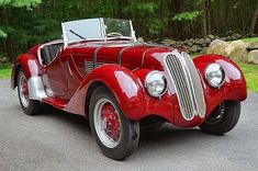 an old red car is parked on the side of the road in front of some trees