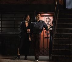 a man and woman standing in front of some stairs at night with the lights on