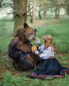 a woman sitting on the ground next to a brown bear and eating food from a basket