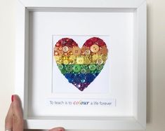 a person holding up a white frame with a heart made out of buttons in the shape of a rainbow