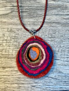 a multicolored felt pendant hangs from a leather cord on a wooden surface,