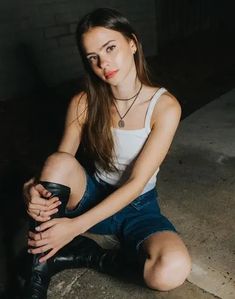 a young woman sitting on the ground with her legs crossed, wearing black boots and a white tank top