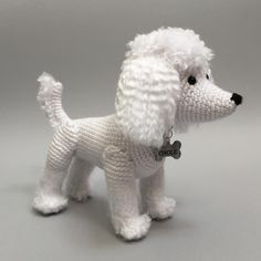 a crocheted white poodle dog with a bone in its mouth is standing on a gray surface