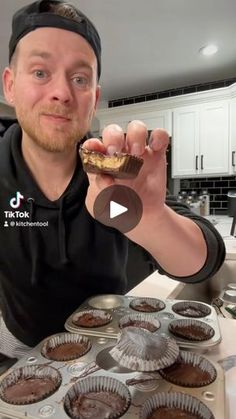 a man holding up a chocolate cupcake in front of some muffin tins