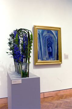 some blue flowers are in a vase on a white pedestal next to a framed painting