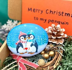 a penguin painted on a rock sitting in a basket with pine cones and christmas decorations