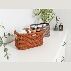 two baskets on top of a white counter next to a potted plant and candles