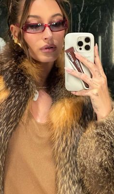 a woman taking a selfie in front of a mirror while wearing sunglasses and a fur coat