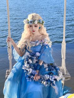 a woman in a blue dress is sitting on a swing by the water with flowers