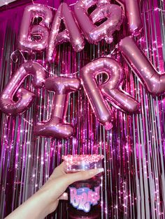 a person holding a drink in front of pink balloons and streamers that spell out the word baby girl