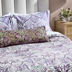 a bed with purple and green pillows on top of it next to a night stand