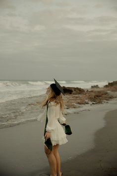 a woman in a white dress and black hat walking on the beach near the ocean