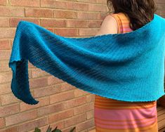 a woman wearing a blue shawl standing in front of a brick wall with her arms wrapped up