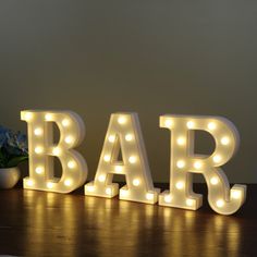 the word bar is lit up with light bulbs on it's sides and sits next to a potted plant