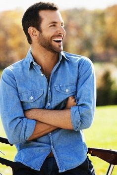 a man with his arms crossed sitting on a bench in a park smiling at the camera