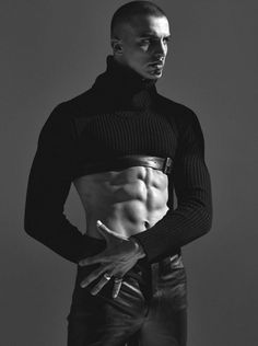 a man with no shirt standing in front of a gray background wearing jeans and a turtle neck sweater