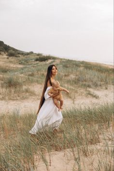 a woman holding a baby in her arms while walking through tall grass on the beach