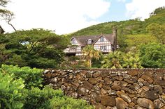 a stone wall in front of a house with trees on the hill side behind it