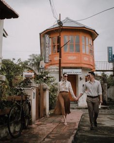 a man and woman holding hands while walking down the street in front of an orange building