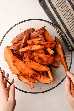 two hands holding a wooden spoon over a bowl full of cooked potato wedges on a white table