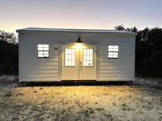 a small white building sitting on top of a dry grass field at dusk with the sun shining through the windows