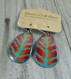 two pairs of earrings with blue and red leaves on them sitting on a wooden table