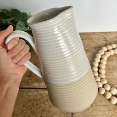 a hand holding a white ceramic vase on top of a wooden table next to a beaded necklace