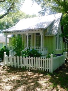 a small green house with a white picket fence and trees in the front yard on a sunny day