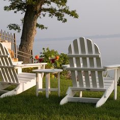 two white adironda chairs sitting in the grass next to a tree and water