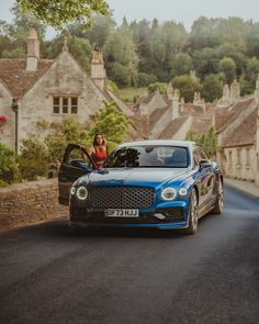 a woman sitting in the open door of a blue car on a road with houses behind her