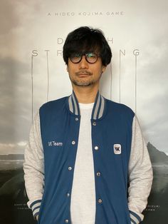 a man standing in front of a poster wearing glasses and a blue jacket with white sleeves